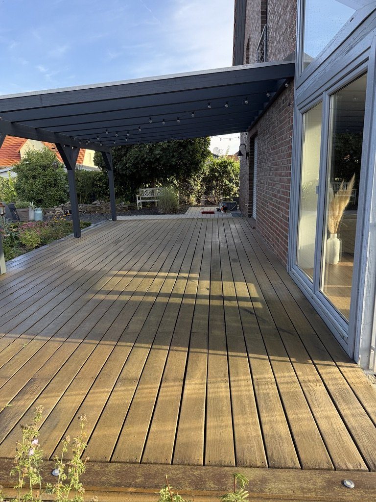 Holzterrasse unter der Pergola vor der Reinigung, das Pflaster grau, der Schachtdeckel mit Mosaik kaum zu sehen