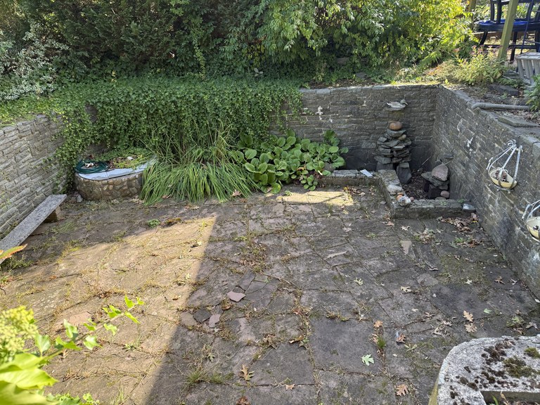 Eingelassener Sitzplatz vor der Arbeit, Unkraut in den Fugen und Bewuchs über der Mauer